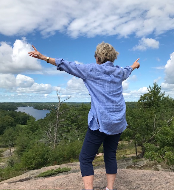 Margaret Anne Cutler standing triumphantly with arms wide open atop a rocky hill overlooking a lake in Muskoka, embodying the joy, vitality and active lifestyle she has maintained well into her 80s
