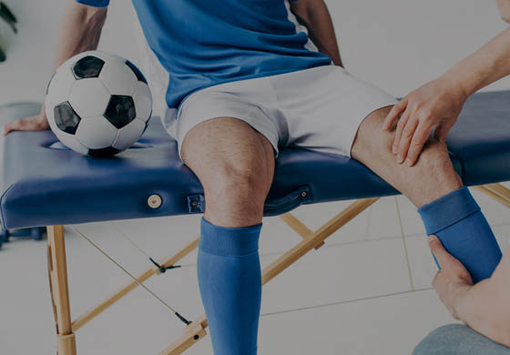 Young male soccer playing sitting on a table, with a physiotherapist's hands working on his knee